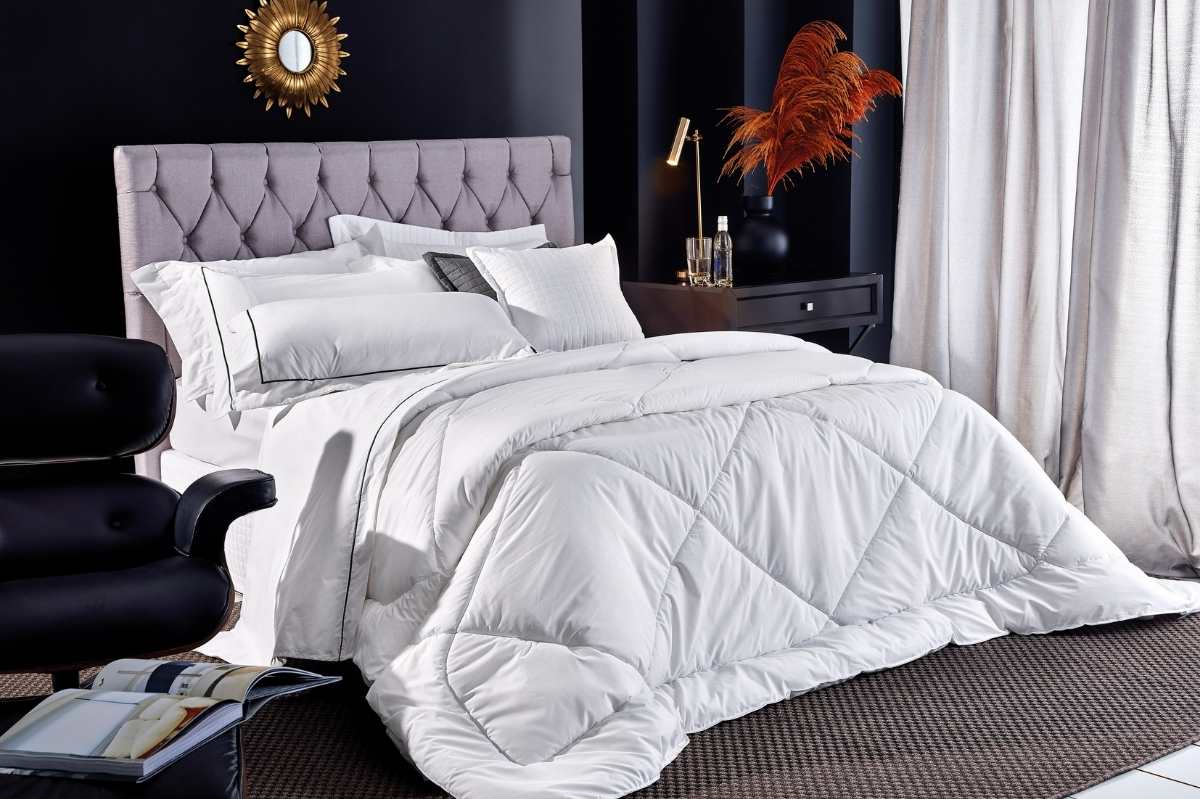 Cama de hotel com edredom branco tipo duvet, almofadas decorativas e cabeceira estofada em tom claro. O ambiente elegante e bem iluminado transmite conforto, limpeza e sofisticação típicos de hospedagens profissionais.