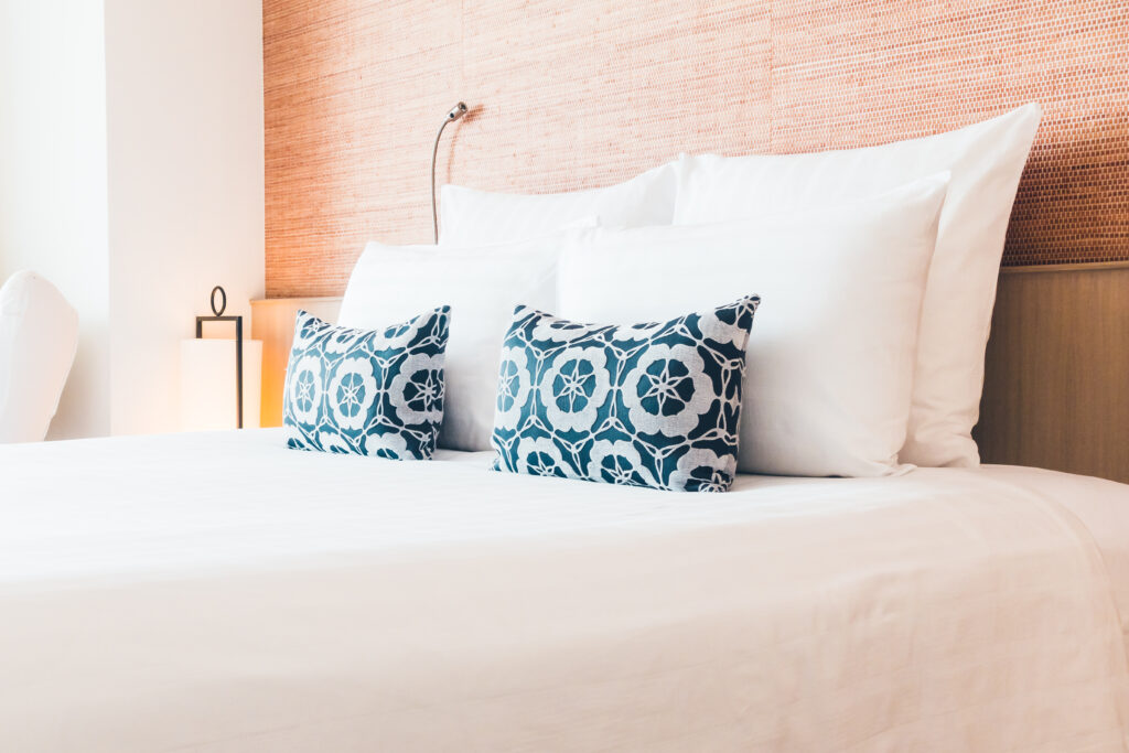 Cama de hotel com enxoval branco e duas almofadas decorativas azuis com estampa geométrica.