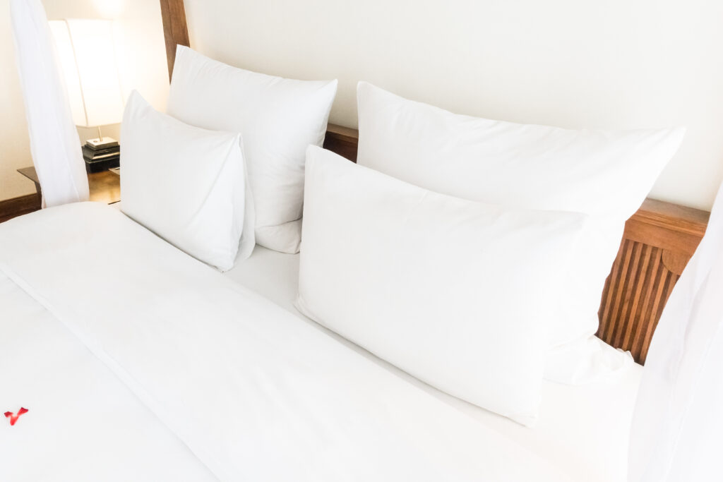 Close de cama de hotel com lençóis brancos e vários travesseiros, cabeceira de madeira e abajur.