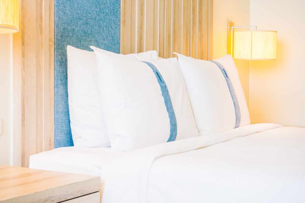 Quarto iluminado com cama de hotel, travesseiros brancos com faixa azul e luminária de parede amarela.