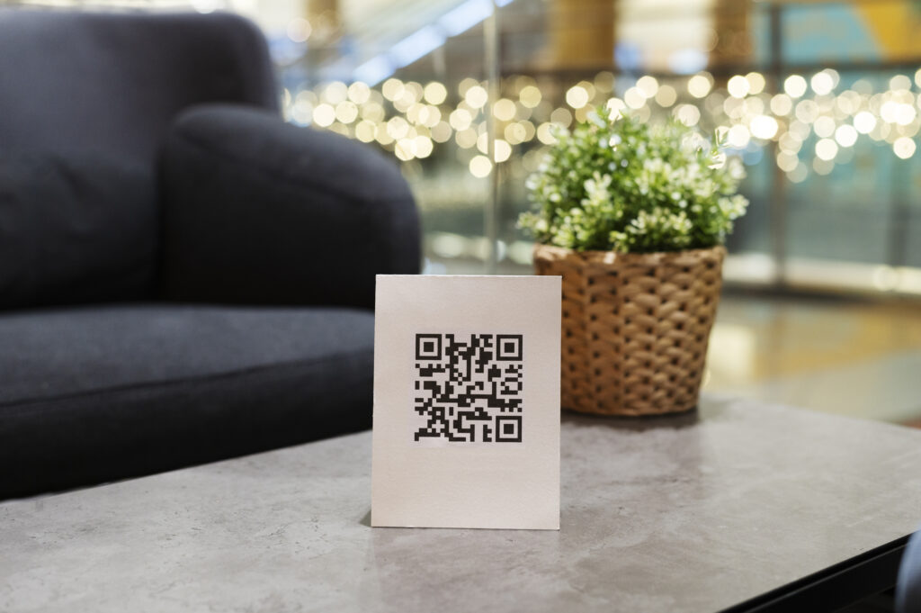 Cartão com código QR sobre mesa de centro em área de convivência de hotel, ao lado de um vaso decorativo. A foto simboliza praticidade e modernização nos serviços de hospedagem, como acesso digital a informações ou regras para hóspedes.