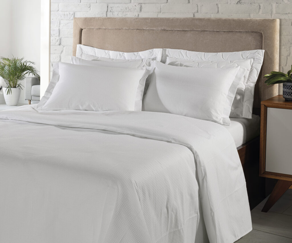 Cama de hotel com enxoval branco completo, composta por lençol, fronhas e colcha em tecido texturizado. O ambiente claro e moderno, com cabeceira estofada e plantas decorativas, transmite conforto, elegância e sensação de limpeza.