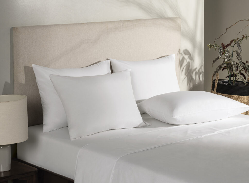 Cama de hotel com enxoval branco completo, composta por lençol, fronhas e travesseiros dispostos de forma organizada. O ambiente iluminado e minimalista transmite sensação de conforto, limpeza e sofisticação típica de hospedagens profissionais.