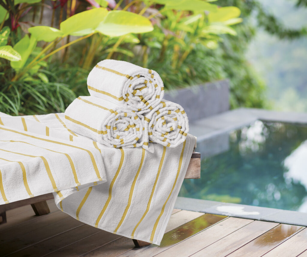 Toalhas de piscina brancas com listras amarelas dobradas sobre espreguiçadeira à beira da piscina. O ambiente tropical com plantas ao redor transmite sensação de frescor, conforto e hospitalidade, ideal para hotéis e pousadas.