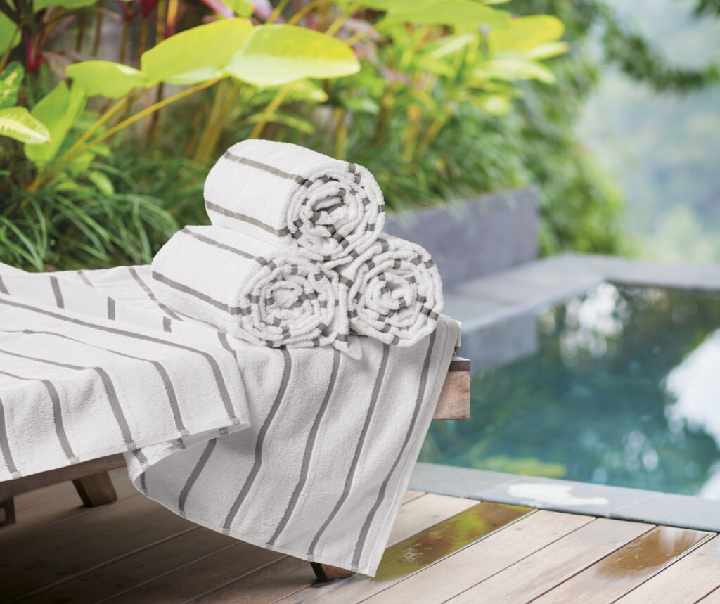 Toalhas de piscina brancas com listras cinza dobradas e dispostas sobre espreguiçadeira à beira da piscina. A cena transmite sensação de conforto, limpeza e elegância, representando o cuidado com o enxoval profissional em áreas de lazer de hotéis e pousadas.