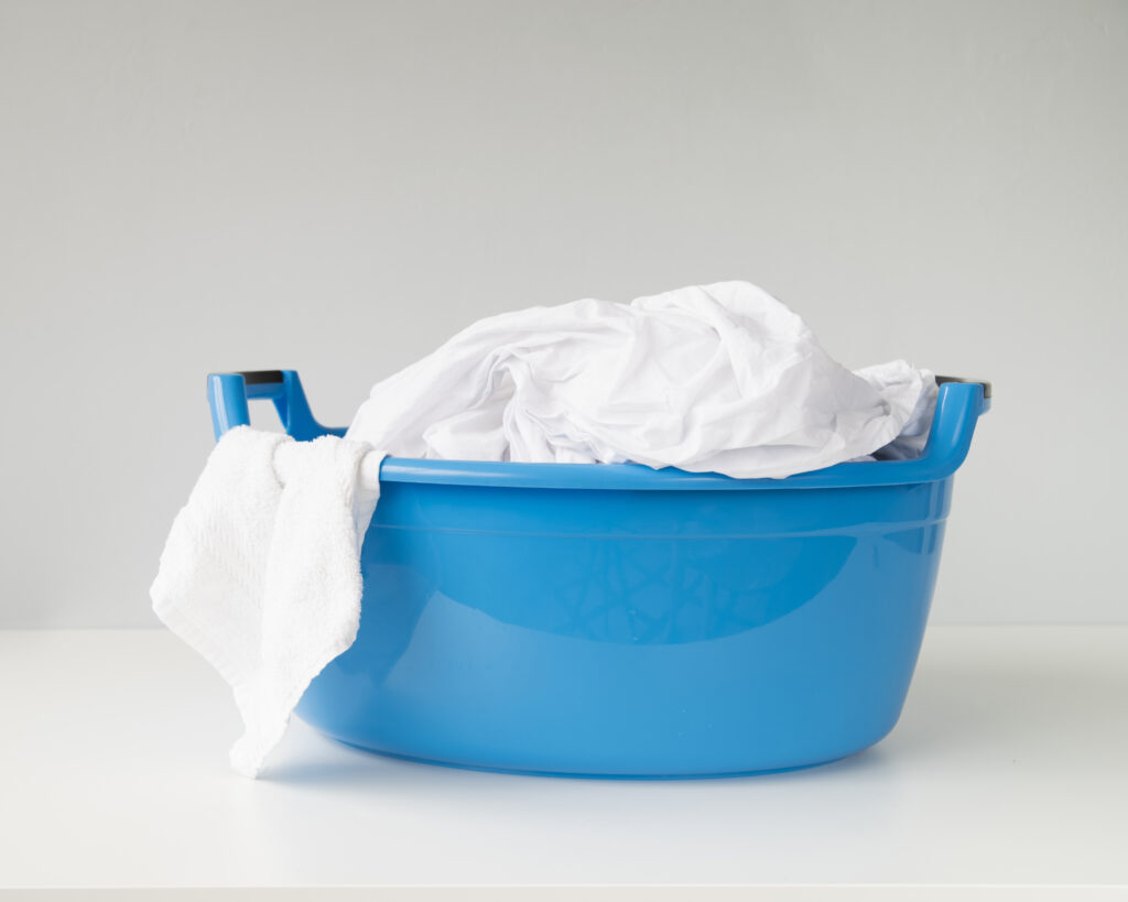 Bacia plástica azul sobre superfície branca, contendo lençóis e toalhas brancas limpas, com fundo neutro.