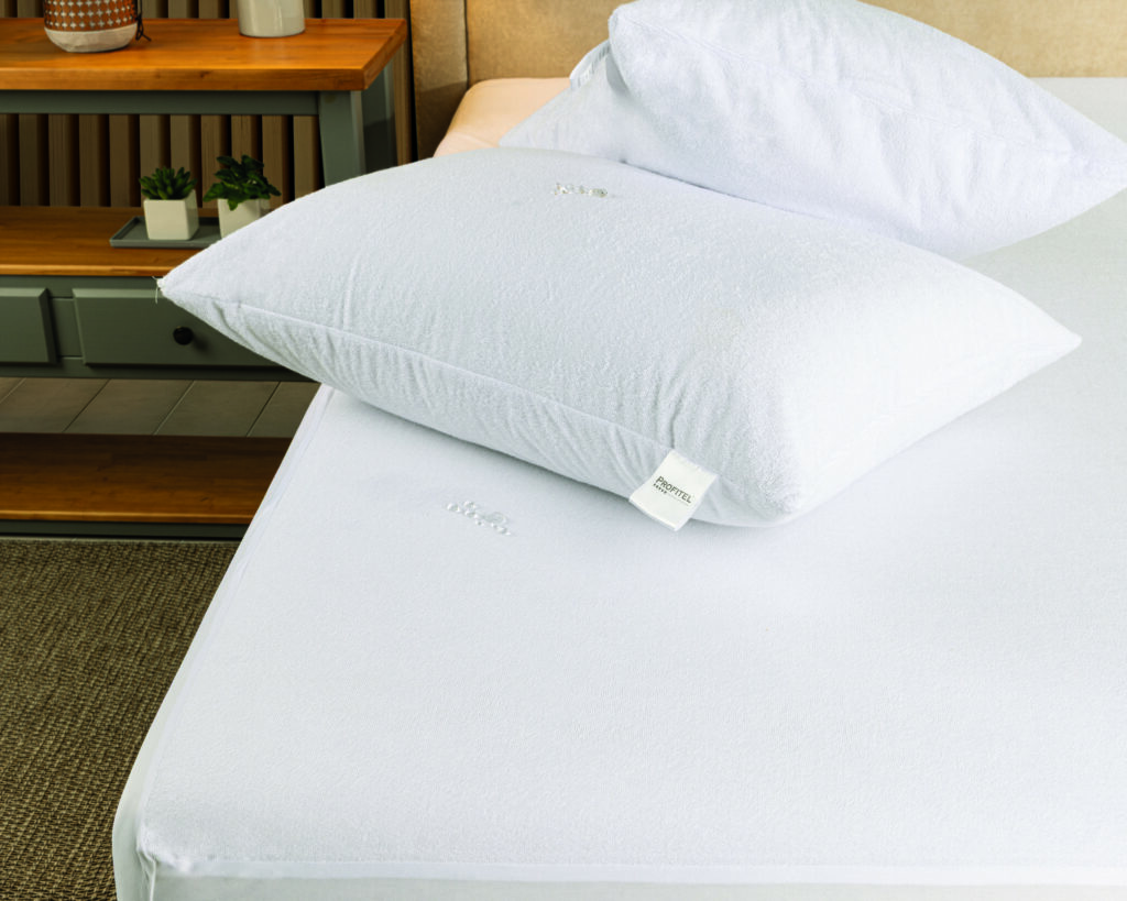 Protetor de travesseiro atoalhado branco sobre colchão com capa impermeável; gotas d’água evidenciam a barreira contra líquidos. Enxoval profissional.
