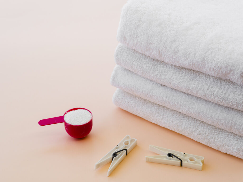 Três toalhas brancas empilhadas, ao lado de uma colher medidora com sabão em pó vermelho e dois prendedores de roupa brancos sobre fundo pêssego claro.