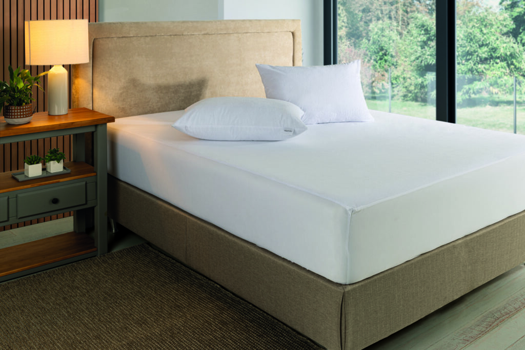 Quarto com cama box e cabeceira bege; protetor de colchão atoalhado branco ajustado e dois travesseiros, mesa lateral com luminária e plantas. Enxoval profissional.