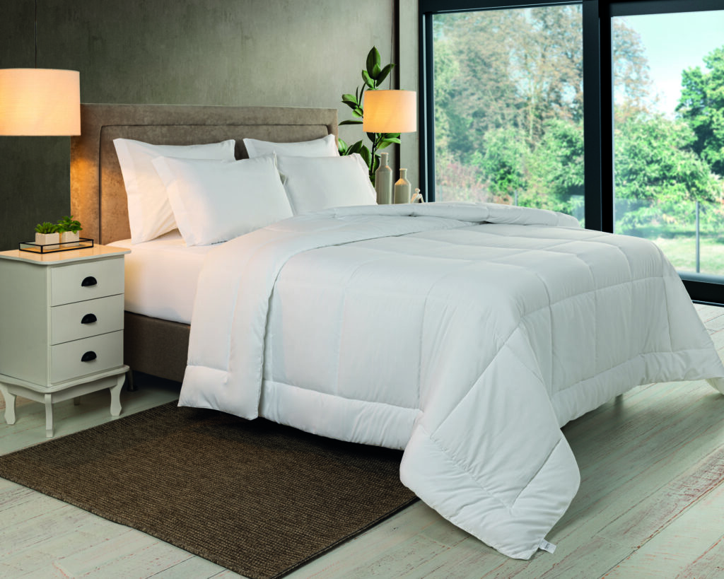 Quarto iluminado com cama arrumada em enxoval branco, edredom volumoso e travesseiros alinhados, ao lado de criado-mudo claro e grandes janelas com vista para área verde.