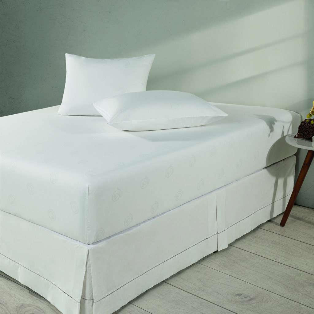 Cama box com lençol branco ajustado de padrão discreto e dois travesseiros, saia para box e mesa lateral, em ambiente minimalista com luz natural. Enxoval profissional para hotelaria.