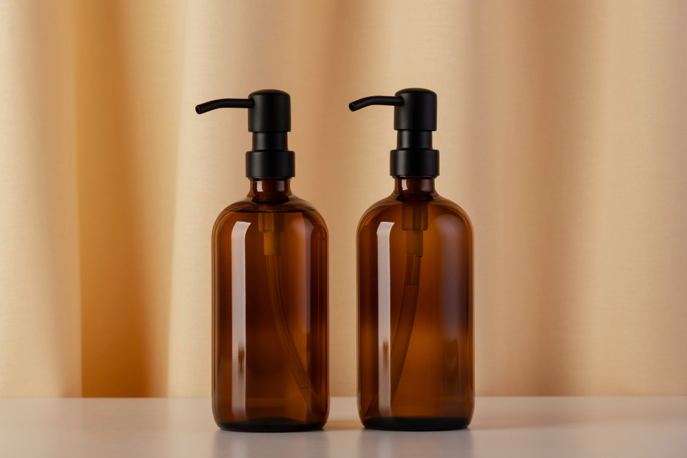 Dois frascos de vidro âmbar com válvula pump preta posicionados lado a lado sobre uma superfície clara, com fundo em tom bege.
