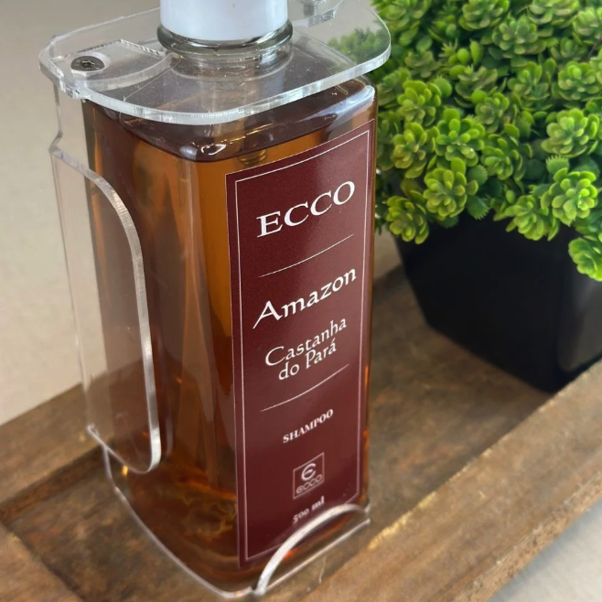 Frasco de shampoo Ecco Amazon Castanha do Pará em suporte transparente sobre base de madeira, com planta decorativa verde ao fundo.