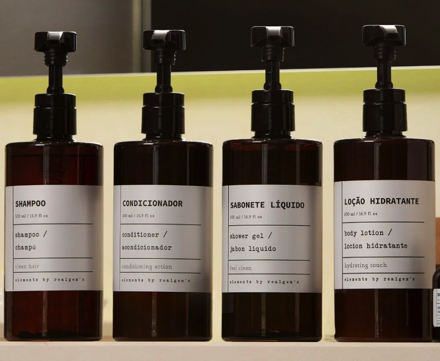 Quatro frascos âmbar com válvula pump preta alinhados em uma prateleira, rotulados como shampoo, condicionador, sabonete líquido e loção hidratante.