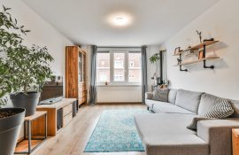 Ambiente residencial moderno com sofá cinza, móveis de madeira, tapete azul e janela ampla com cortinas cinzas.