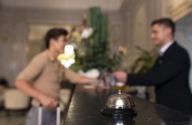 Recepcionista de hotel atendendo hóspede durante o check-in, com um sino de balcão em destaque. A imagem representa o acolhimento e a atenção nos primeiros momentos da hospedagem.