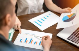 Pessoas reunidas em mesa de trabalho analisando relatórios com gráficos ao lado de um notebook, em reunião de análise de resultados.