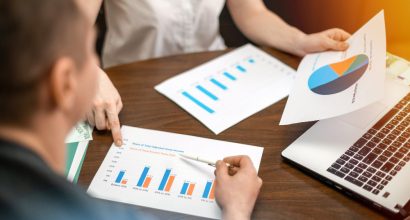 Pessoas reunidas em mesa de trabalho analisando relatórios com gráficos ao lado de um notebook, em reunião de análise de resultados.