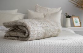 Manta bege dobrada sobre uma cama arrumada com roupas de cama claras, em um quarto decorado em tons neutros com detalhes minimalistas ao fundo.