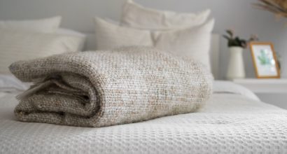 Manta bege dobrada sobre uma cama arrumada com roupas de cama claras, em um quarto decorado em tons neutros com detalhes minimalistas ao fundo.