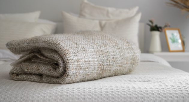 Manta bege dobrada sobre uma cama arrumada com roupas de cama claras, em um quarto decorado em tons neutros com detalhes minimalistas ao fundo.