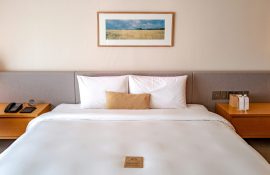 Cama de hotel com enxoval branco, dois travesseiros e almofada bege; cabeceira estofada cinza, quadro de paisagem e mesas laterais com telefone e amenities.