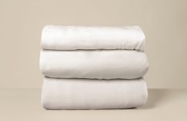 Três jogos de cama brancos dobrados e empilhados, exibidos sobre fundo neutro.