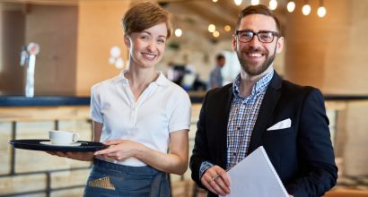 Funcionária de hotel com bandeja e xícara de café ao lado de um homem com prancheta, sorrindo em um lobby com luzes pendentes ao fundo.