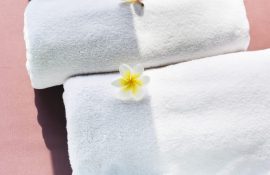 Duas toalhas brancas felpudas enroladas, decoradas com flor branca e amarela sobre superfície rosa.