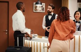 Casal realizando check-in em hotel, sendo atendido com cordialidade por recepcionistas uniformizados. A cena transmite hospitalidade e atenção aos detalhes, reforçando a importância do bom atendimento na experiência do hóspede.