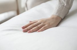 Mão com unhas claras tocando um lençol branco, destacando a maciez e o conforto do enxoval profissional de cama.