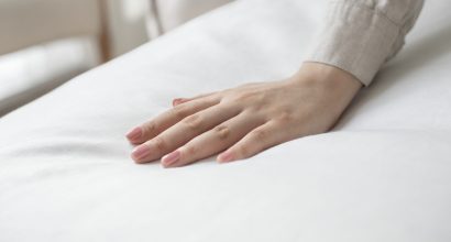 Mão com unhas claras tocando um lençol branco, destacando a maciez e o conforto do enxoval profissional de cama.
