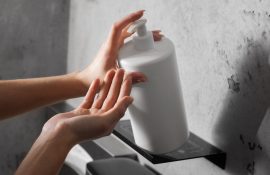 Mãos acionando o pump de um frasco branco de dispenser apoiado em uma prateleira de banheiro com parede cinza.