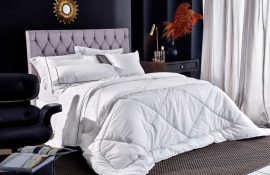 Cama de hotel com edredom branco tipo duvet, almofadas decorativas e cabeceira estofada em tom claro. O ambiente elegante e bem iluminado transmite conforto, limpeza e sofisticação típicos de hospedagens profissionais.