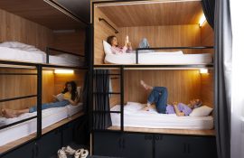 Dormitório coletivo moderno com quatro cápsulas; hóspedes relaxam em três delas; roupa de cama branca e armários numerados.