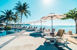 Área de piscina de hotel à beira-mar com espreguiçadeiras brancas, guarda-sóis e palmeiras sob céu ensolarado. A imagem transmite clima tropical, conforto e lazer, ideal para ilustrar ambientes de alta temporada em hospedagens profissionais.