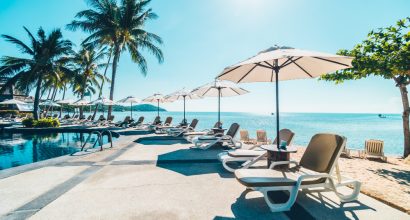 Área de piscina de hotel à beira-mar com espreguiçadeiras brancas, guarda-sóis e palmeiras sob céu ensolarado. A imagem transmite clima tropical, conforto e lazer, ideal para ilustrar ambientes de alta temporada em hospedagens profissionais.