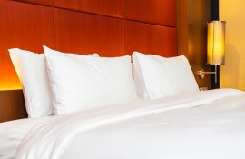 Detalhe de cama arrumada com enxoval branco e iluminação aconchegante ao lado da cabeceira.